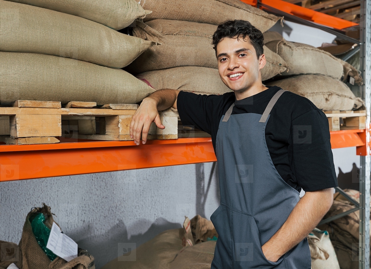 Portrait of a young male barista in an apron leaning on a shelf in a warehouse Warehouse worker posing in front of a camera