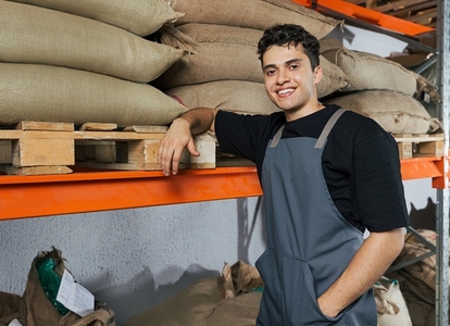 Portrait of a young male barista in an apron leaning on a shelf in a warehouse Warehouse worker posing in front of a camera Portrait of a young male barista in an apron leaning on a shelf in a warehouse Warehouse worker posing in front of a camera